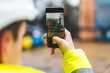 © LOOKSLIKEPHOTO.COM - Architect holding a smartphone on construction site - young construction worker is using mobile phone on site - Construction worker with building plans and cellphone - Focus on mobile. warm filter