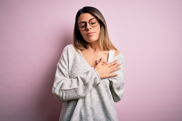 Sticker - Young beautiful brunette woman wearing casual sweater and glasses over pink background smiling with hands on chest with closed eyes and grateful gesture on face. Health concept.