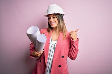 Sticker - Young beautiful brunette architect woman wearing safety helmet holding blueprints pointing and showing with thumb up to the side with happy face smiling