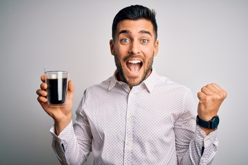 Young handsome man drinking a cup of hot coffee over white isolated background screaming proud and celebrating victory and success very excited, cheering emotion