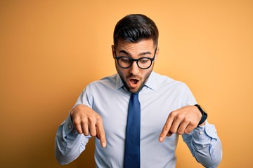 Sticker - Young handsome businessman wearing tie and glasses standing over yellow background Pointing down with fingers showing advertisement, surprised face and open mouth