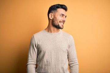 Wall Mural - Young handsome man wearing casual sweater standing over isolated yellow background looking away to side with smile on face, natural expression. Laughing confident.