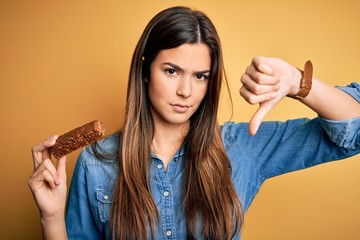 Poster - Young beautiful girl holding healthy protein bar standing over isolated yellow background with angry face, negative sign showing dislike with thumbs down, rejection concept