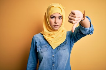 Poster - Young beautiful girl wearing muslim hijab standing over isolated yellow background looking unhappy and angry showing rejection and negative with thumbs down gesture. Bad expression.