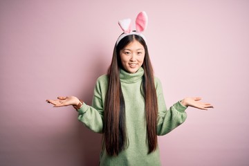 Canvas Print - Young asian woman wearing cute easter bunny ears over pink background clueless and confused expression with arms and hands raised. Doubt concept.