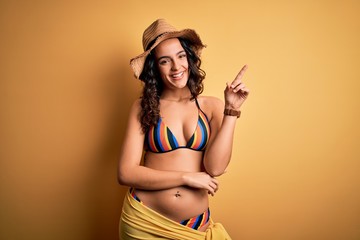 Young beautiful woman with curly hair on vacation wearing bikini and summer hat with a big smile on face, pointing with hand and finger to the side looking at the camera.