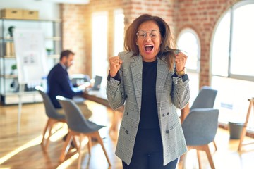 Middle age beautiful businesswoman wearing jacket and glasses standing at the office celebrating surprised and amazed for success with arms raised and open eyes. Winner concept.