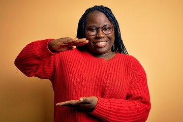 Sticker - African american plus size woman with braids wearing casual sweater over yellow background gesturing with hands showing big and large size sign, measure symbol. Smiling looking at the camera. Measure