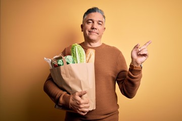 Sticker - Middle age handsome grey-haired man holding paper bag with food over yellow background very happy pointing with hand and finger to the side