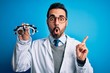 Young handsome optical man with beard holding optometry glasses over blue background Surprised pointing with finger to the side, open mouth amazed expression.
