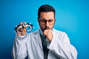 Sticker - Young handsome optical man with beard holding optometry glasses over blue background feeling unwell and coughing as symptom for cold or bronchitis. Health care concept.