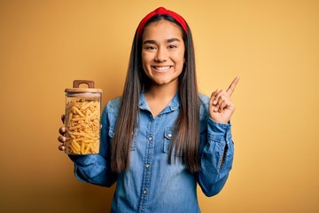 Canvas Print - Young beautiful asian woman holding jar of Italian dry pasta macaroni over yellow background very happy pointing with hand and finger to the side