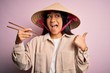 © Krakenimages.com - Young thai woman wearing traditional conical asian hat eating food using chopsticks pointing and showing with thumb up to the side with happy face smiling