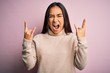 © Krakenimages.com - Young beautiful asian woman wearing casual turtleneck sweater over pink background shouting with crazy expression doing rock symbol with hands up. Music star. Heavy concept.