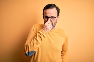 Wall Mural - Middle age hoary man wearing casual sweater and glasses over isolated yellow background smelling something stinky and disgusting, intolerable smell, holding breath with fingers on nose. Bad smell