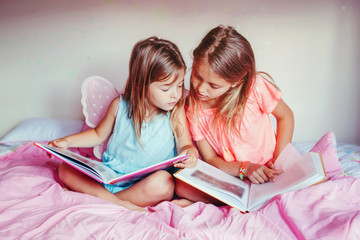 Happy Caucasian sisters girls reading books in the bedroom. Children siblings at home spending time together. Kids education development learning. Real people authentic lifestyle.