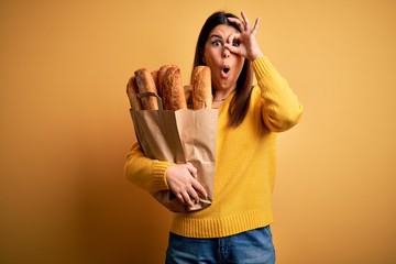 Poster - Young beautiful woman holding a bag of fresh healthy bread over yellow background doing ok gesture shocked with surprised face, eye looking through fingers. Unbelieving expression.