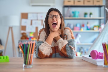 Poster - Young beautiful teacher woman wearing sweater and glasses sitting on desk at kindergarten shouting suffocate because painful strangle. Health problem. Asphyxiate and suicide concept.