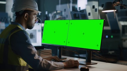 Leinwandbilder - Engineer Wearing Safety Vest and Hardhat Works on a Computer, Two Monitor Screens Show Chroma Key / Green Screen Displays. Modern High-Tech Industry 4.0 Electronics Production Factory