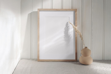 Portrait empty wooden frame mockup with modern ceramic vase, dry grass in sunlight, long shadows. White beadboard wainscot wall paneling background. Scandinavian interior, home design. Art concept.