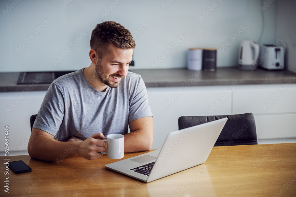 Young smiling man dressed casual sitting at dining table, holding mug with morning coffee and looking at laptop. He is visiting sites for online dating. - obrazy, fototapety, plakaty 