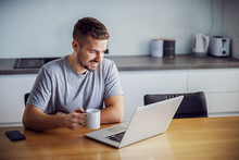 Young Smiling Man Dressed Casual Sitting At Dining Table, Holding Mug With Morning Coffee And Looking At Laptop. He Is Visiting Sites For Online Dating.