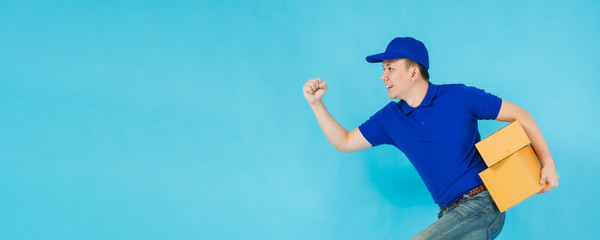 Wall Mural - Asian happy delivery man wearing a blue shirt running and carrying paper parcel boxes isolated on on blue colour background with copy space.Concept of Postal delivery service.