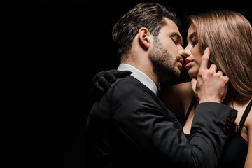 side view of handsome man in suit kissing attractive woman isolated on black