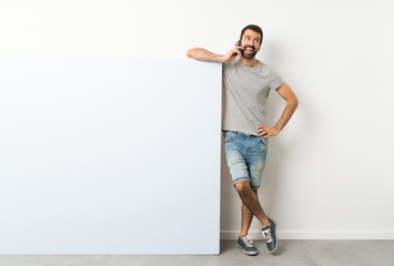 Wall Mural - Young handsome man with beard holding a big blue empty placard keeping a conversation with the mobile phone