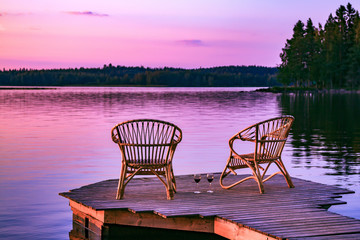 Two rattan chairs and glasses of red wine on a pier overlooking a lake at sunset in Finland