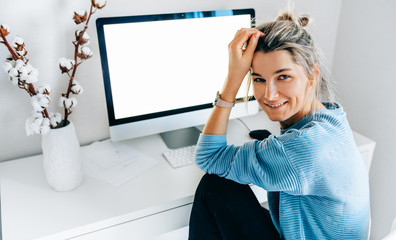 Smiling young freelance businesswoman using desktop pc in the office. The blonde female sits indoors at home working on a computer with a blank screen for your text message or content.