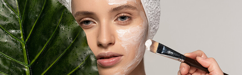 Wall Mural - panoramic shot of beautiful girl applying mask on face with cosmetic brush, isolated on grey with leaf