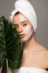 Wall Mural - attractive girl with cosmetic mask on face and towel on head posing with green leaf, isolated on grey