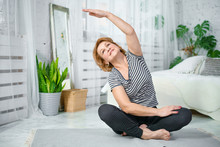 Senior Woman Exercising While Sitting In Lotus Position. Active Mature Woman Doing Stretching Exercise In Living Room At Home.