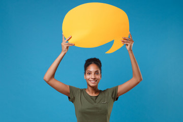 Smiling young african american woman girl in casual t-shirt posing isolated on bright blue background. People lifestyle concept. Mock up copy space. Hold yellow empty blank Say cloud, speech bubble.