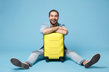 Wall Mural - Laughing traveler tourist man in summer yellow casual clothes isolated on blue wall background. Male passenger traveling abroad to travel on weekend. Air flight journey concept. Sitting near suitcase.