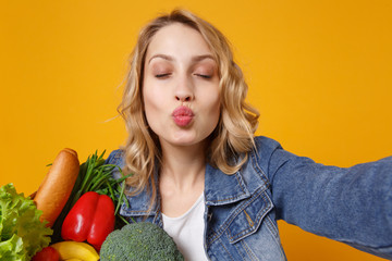 Wall Mural - Close up of young woman in denim clothes isolated on orange background. Delivery service from shop or restaurant concept. Hold bag for takeaway with food products doing selfie shot on mobile phone.