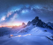 Milky Way over snowy mountains and blurred car headlights on the winding road at night in winter. Beautiful landscape with starry sky, snow covered rocks, house, roadway at sunset. Space and galaxy