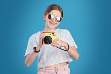 Portrait Of Positive Teenage Girl In Sunglasses Photographing On Instant Camera Against Blue Background