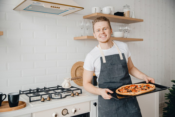 European male chef smiles and holds baking tray with hot Italian pizza in bright kitchen