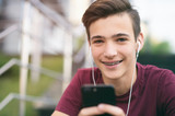 Close-up portrait of a smiling young man with a smartphone, in the street.  Happy teenage boy is using mobile phone, outdoors. Cheerful teenager spends time in social networks using cell phone.