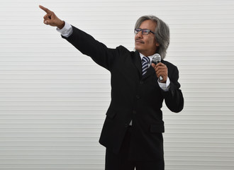 Happy elderly asian businessman in black suit and eyeglasses speaking with microphone and pointing finger up for the future standing over white wall background, Business presentation and seminar conce