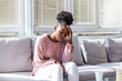 © Graphicroyalty - Portrait of a young black girl sitting on the couch at home with a headache and back pain. Beautiful woman suffering from chronic daily headaches. Sad woman holding her head because sinus pain