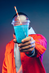 Wall Mural - Vertical picture of young arabian man holding plastic cup with blue cocktail in it. Drinking straw in between. Guy wear red jacket. Isolated over colorful background.