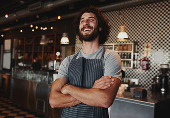 Happy young man wearing apron standing with folded arms in coffee house