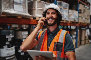 Wall Mural - Warehouse worker using digital tablet while in conversation over mobile phone in warehouse wearing white helmet and safety vest