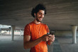 © StratfordProductions - Portrait of a smiling young male runner standing under the concrete bridge with earphone in his ears typing on a smart phone