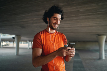 portrait of a smiling young male runner standing under the concrete bridge with earphone in his ears