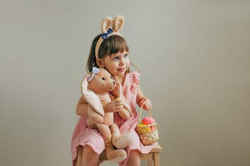 Wall Mural - Little girl with bunny ears with bunny toy and Easter painted eggs on beige background with copy space