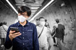 © zephyr_p - Asian man wearing surgical face mask using smartphone in subway tunnel with crowded people walking. Wuhan coronavirus (COVID-19) outbreak prevention in public area. Health care and medical concept